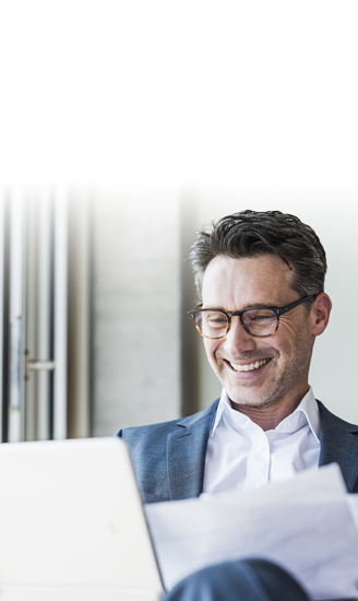 A smiling businessman looks over reports on his laptop as he uses his managed network services from Glo Fiber. The subject is holding papers and reviewing data while using a managed wifi connection.